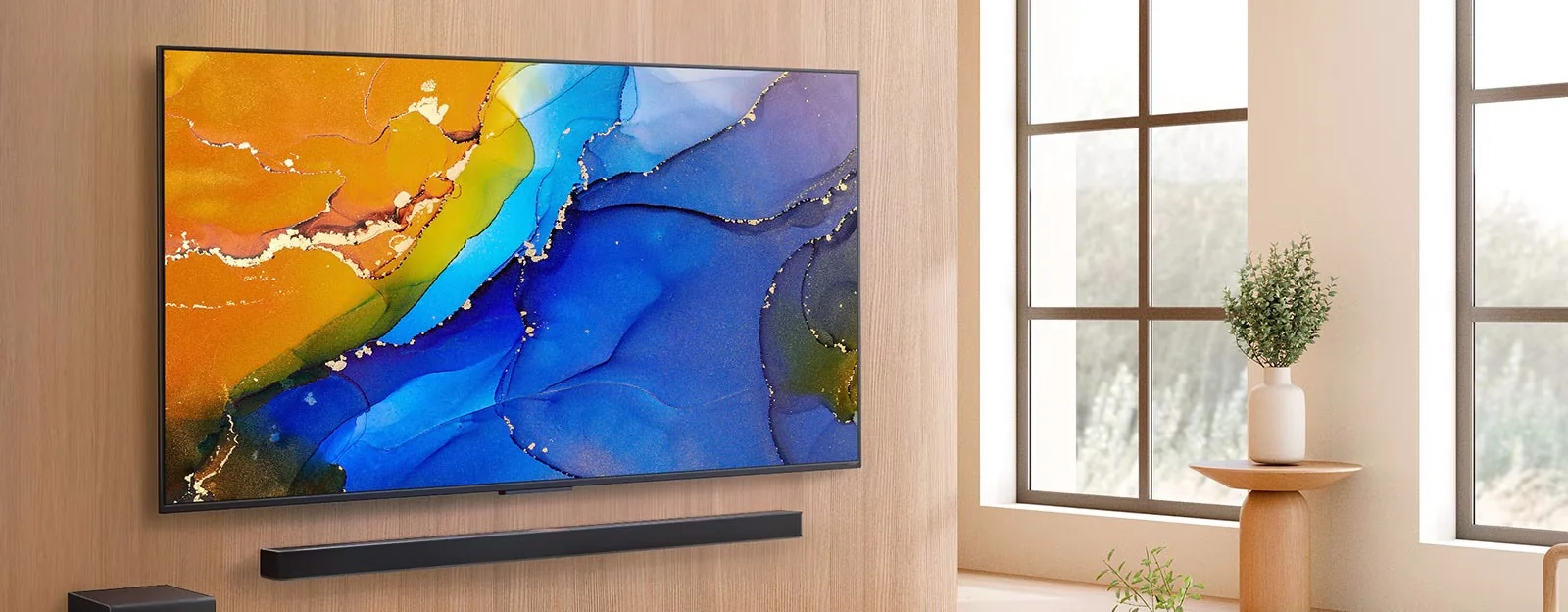 A modern TV mounted on a light wood wall displays vibrant abstract art in blue, orange, and yellow. Below is a black soundbar. A window and plant create a cosy, natural setting.