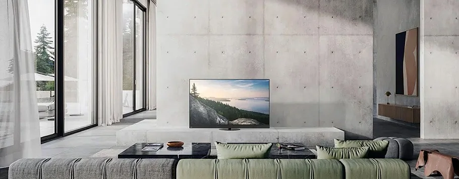 A modern, minimalist living room with large windows showing a forest view. A green sofa faces a flat-screen TV on a concrete wall. The space feels serene and airy.