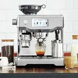 A sleek, silver espresso machine on a wooden counter, brewing coffee into a clear glass. Surrounding are jars of beans, milk, and a silver frothing pitcher, against a white tile backdrop.