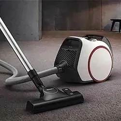 A modern canister vacuum cleaner with a sleek white and black design is on a carpeted floor in a minimalist room, conveying efficiency and style.