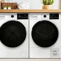 Front view of a modern washer and dryer set with digital displays, set under a wooden countertop. Towels and a potted plant add a homely touch.