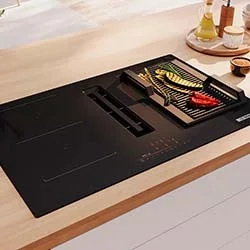 Modern kitchen stove top with a sleek black finish featuring a grilling section with bell peppers and zucchini. Nearby are cooking oils and spices.