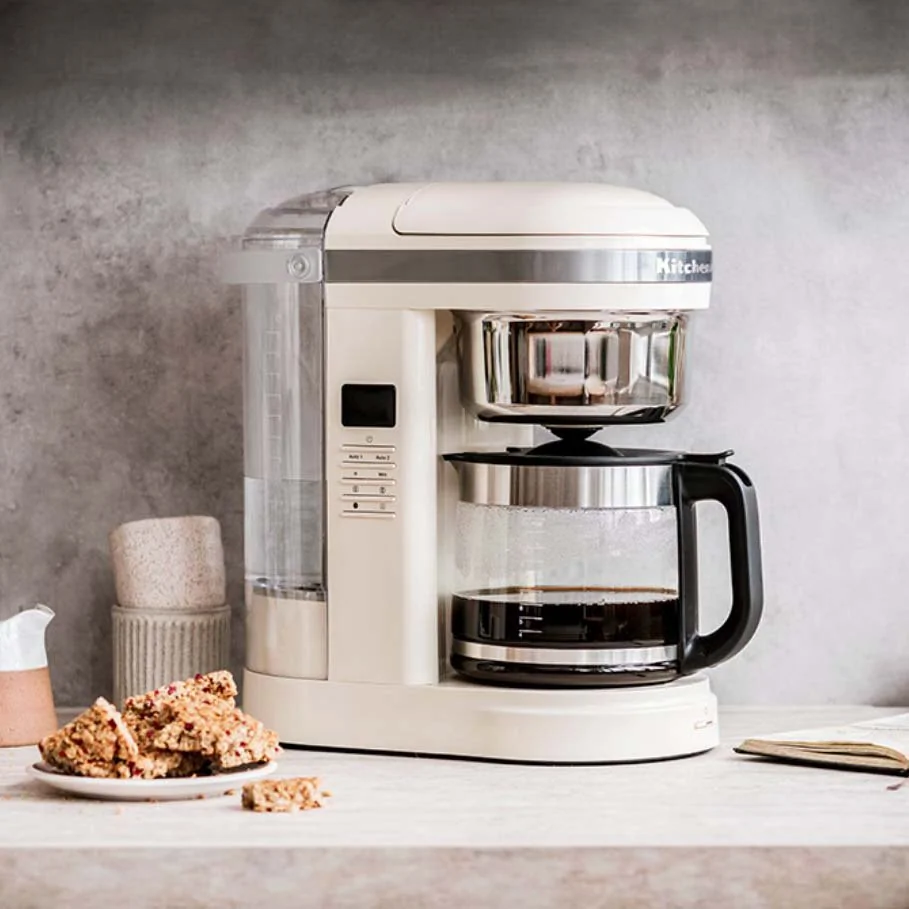 Cream-colored coffee maker brewing fresh coffee, next to a plate of granola squares on a light-coloured countertop.