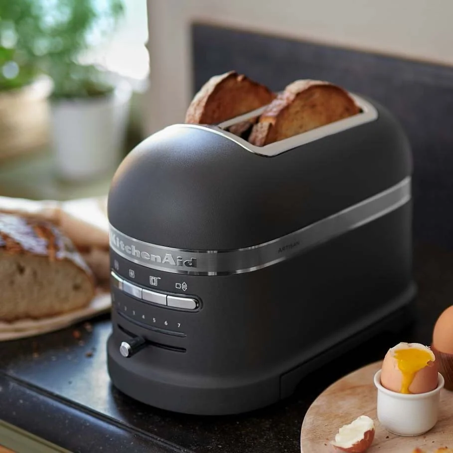 A sleek black toaster with two slices of bread on a kitchen counter, next to a sliced loaf and a soft-boiled egg in an egg cup.