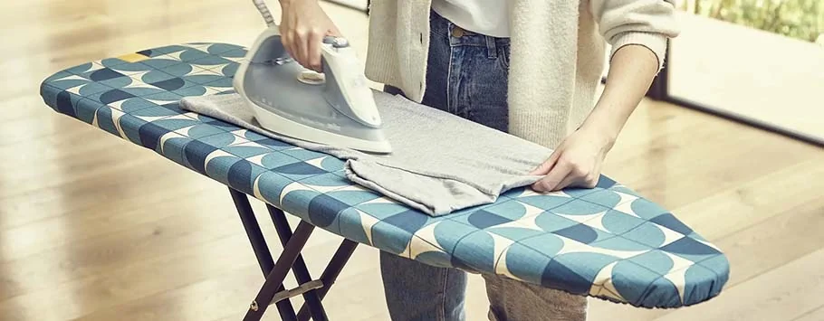A person irons a grey shirt on a patterned blue ironing board in a bright room with hardwood floors, creating a calm, domestic atmosphere.