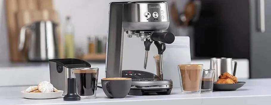 Modern espresso machine on a kitchen counter surrounded by coffee cups, pastries, and a milk frother. The scene conveys a warm, inviting atmosphere.