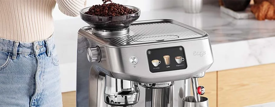 A person in jeans stands beside a sleek silver espresso machine with fresh coffee beans on top. The touchscreen displays drink options, creating a cosy kitchen atmosphere.