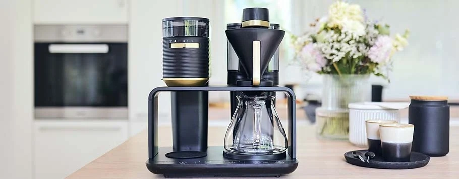 Stylish black and gold coffee maker on a kitchen counter, next to flowers and cups of coffee. The scene is modern and inviting.