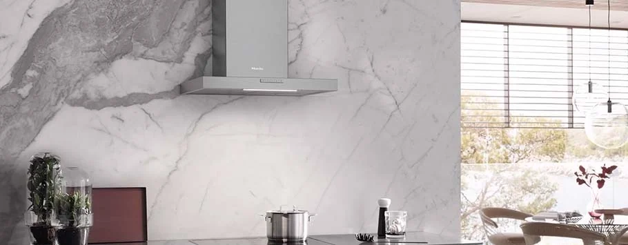 Modern kitchen with a sleek steel range hood on a marbled wall. Minimalist décor includes potted plant, utensils, and a pot. Large windows add natural light.