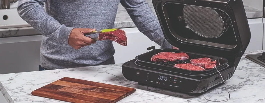 A person places a raw steak into an open indoor grill on a marble countertop. More steaks are inside, and a wooden cutting board is nearby.