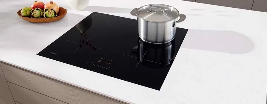 Modern kitchen scene with a sleek black induction cooktop on a white counter. A stainless steel pot is on the burner, and a bowl of vegetables sits nearby.