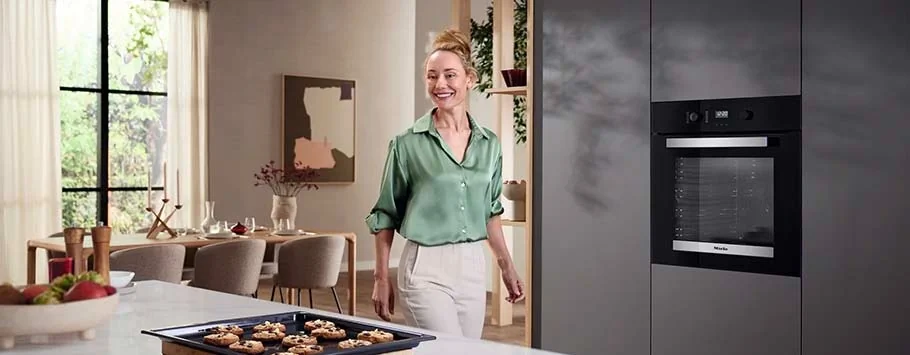Woman in a green blouse smiles in a modern kitchen. Freshly baked cookies are on the counter. Sunlight streams through large windows, creating a bright, cosy atmosphere.