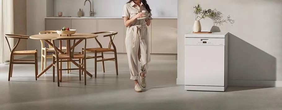 A woman in a beige jumpsuit holds a mug, walking in a modern kitchen with a round wooden table, chairs, and a white dishwasher. The ambiance is calm and minimalist.