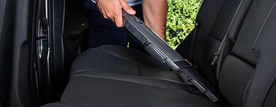 A person uses a handheld vacuum to clean the backseat of a car. The car seats are dark grey, and the background features green foliage. The scene conveys cleanliness and tidiness.