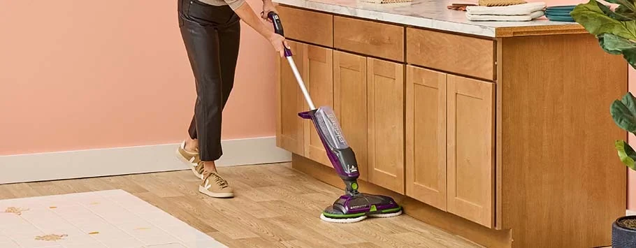 A person cleans a wooden kitchen floor with a purple and white motorized mop. The space is bright and tidy, evoking a sense of cleanliness and efficiency.