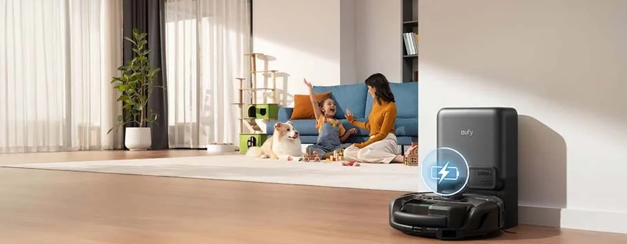 A living room with a robot vacuum charging on hardwood floor. In the background, a woman and child play joyfully with a dog on a white rug.