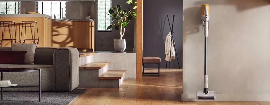 Modern living room with a sleek cordless vacuum mounted on a wall. Features wooden floors, a gray sofa, and bar stools, exuding a clean and cosy feel.