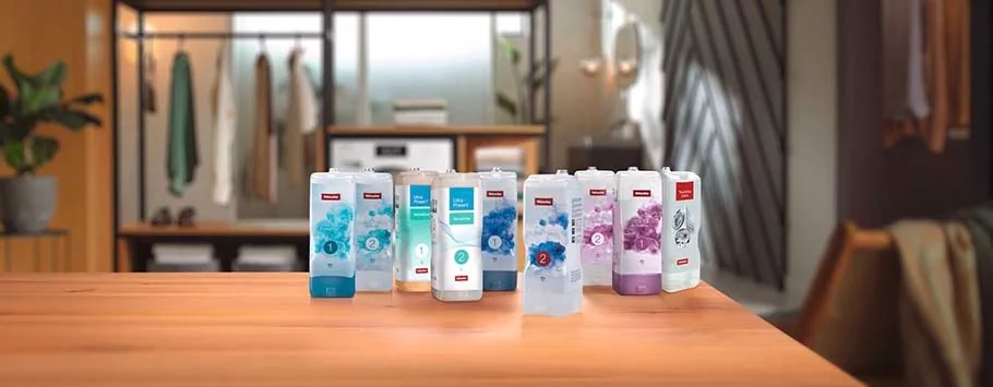 Nine bottles of cleaning products with various coloured labels, including blue and purple, are arranged on a wooden table in a cosy, softly lit room.