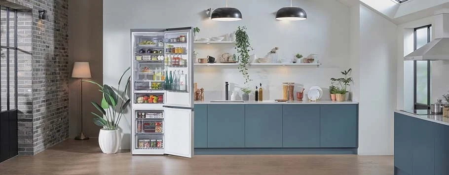 Modern kitchen with open fridge full of organized food, surrounded by blue cabinets and white shelves holding plants and kitchenware, under soft lighting.
