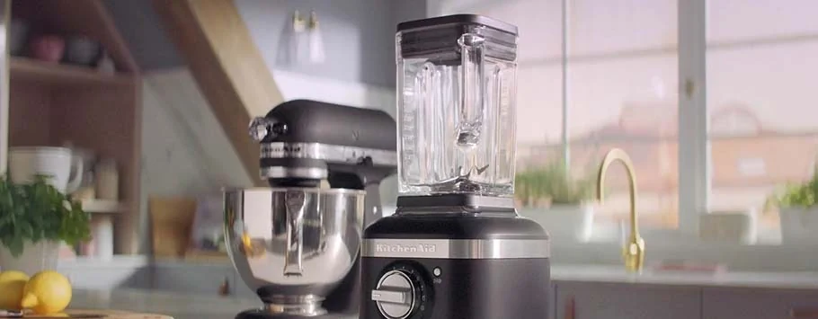 A sleek black KitchenAid blender sits on a modern kitchen counter, next to a black stand mixer. Sunlight streams through a window, creating a warm, inviting atmosphere.