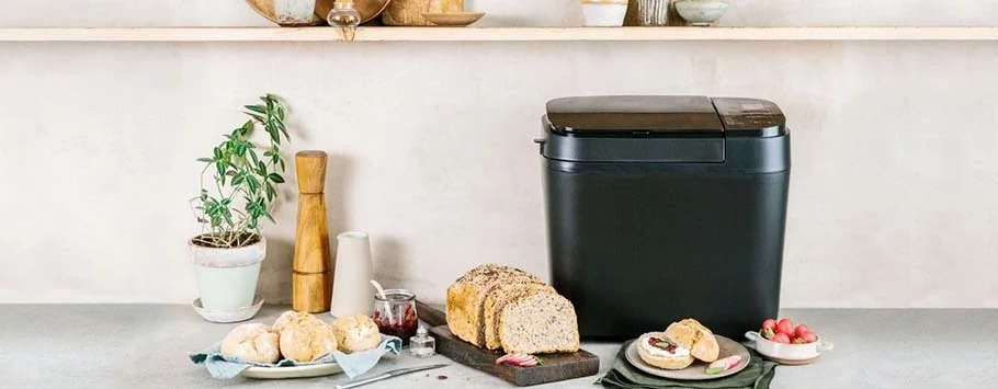 Black compost bin on a kitchen counter with freshly baked bread, rolls, a potted plant, and a pepper mill. The setting is neat and inviting.