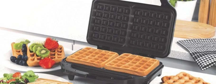 Open waffle maker with two golden waffles inside, surrounded by plates of fruit-topped waffles on a kitchen counter. Fresh and inviting breakfast scene.