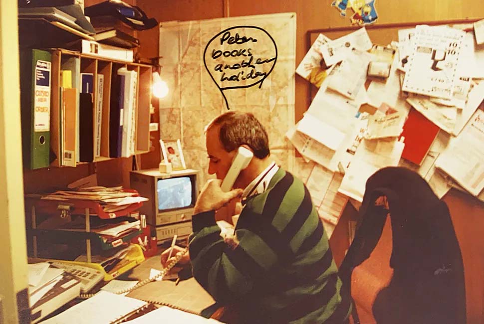 Man in a cluttered office talks on a phone, jotting notes. The wall is filled with papers. A speech bubble humorously reads, "Peter books another holiday."
