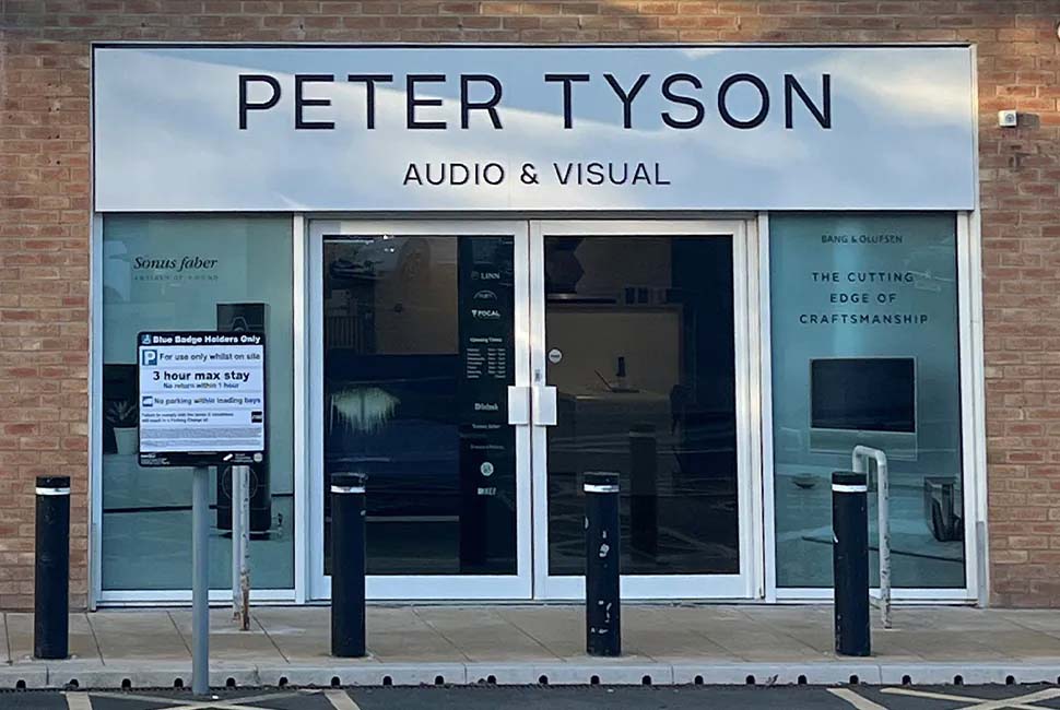 Storefront of "Peter Tyson Audio & Visual" with a brick facade. Large windows display various audio brands. Sign indicates parking rules. Modern, professional tone.
