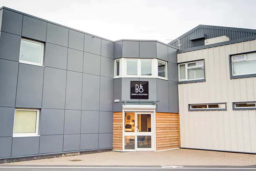 Modern industrial building exterior with grey and beige panels. A large sign above the entrance reads "B&O Bang & Olufsen," conveying a sleek, contemporary vibe.