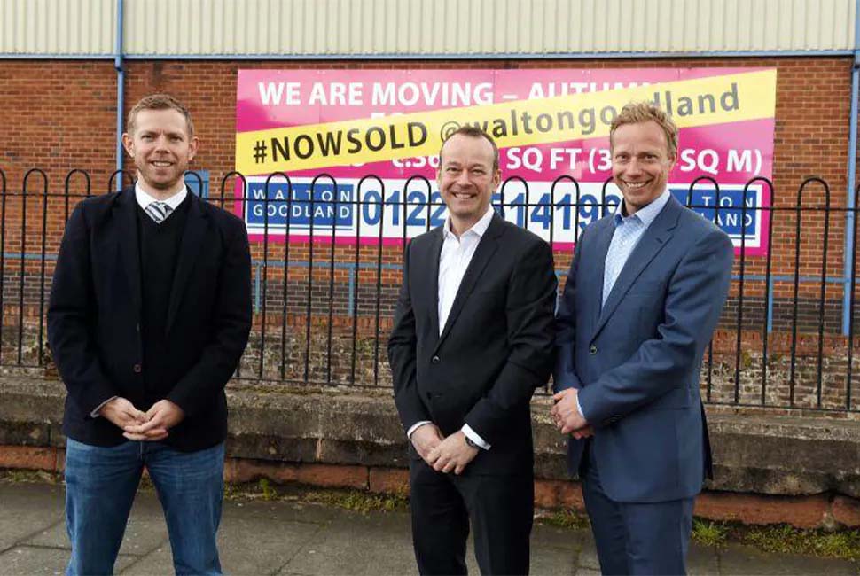 Three smiling men in suits stand in front of a building with a vibrant “We Are Moving” sign. The atmosphere is positive and professional.