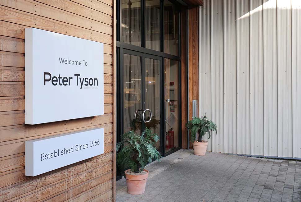 Entrance of a modern building with a large glass door, wooden siding, and a white sign reading "Welcome to Peter Tyson, Established Since 1966." Two potted plants are beside the door.