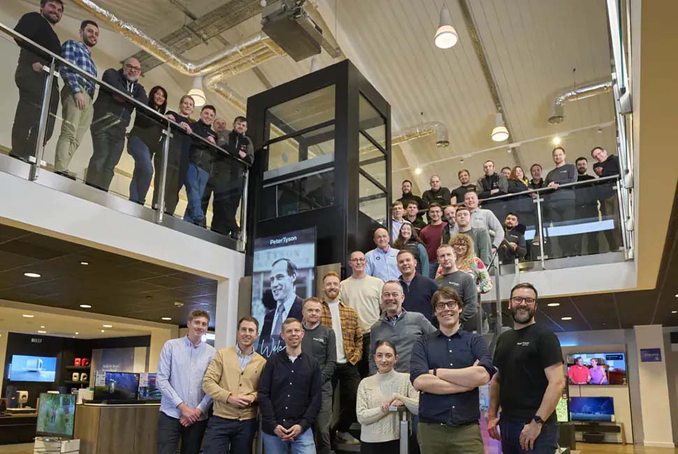 A group of smiling people gathered on two levels of a modern showroom, with sleek electronics on display. The atmosphere is lively and friendly.