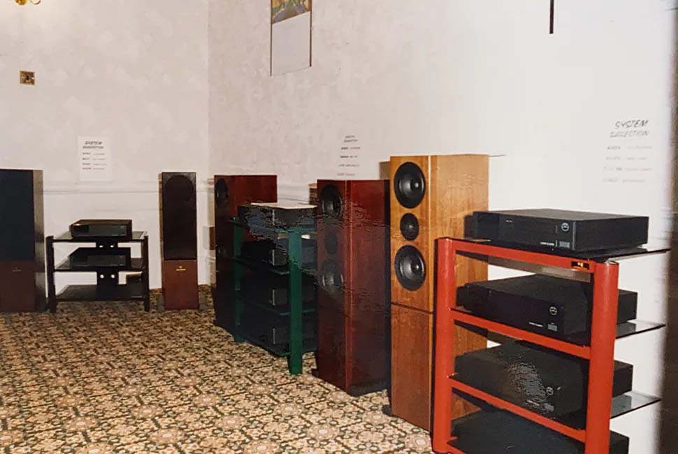 A room features vintage audio equipment with large wooden speakers and shelves holding black stereos. The floral carpet adds a retro touch to the setting.