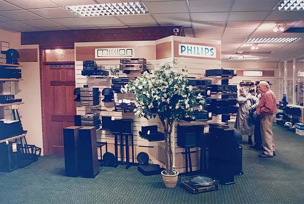 A vintage electronics store with Mission and Philips signs. Audio equipment is displayed around a central potted plant. Two people browse in the background.