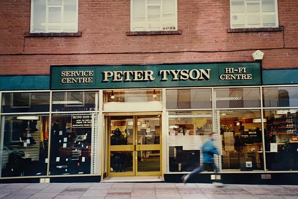 Street view of a hi-fi service center with a green sign reading "Peter Tyson." A person in a blue jacket moves quickly in front, creating a dynamic feel.