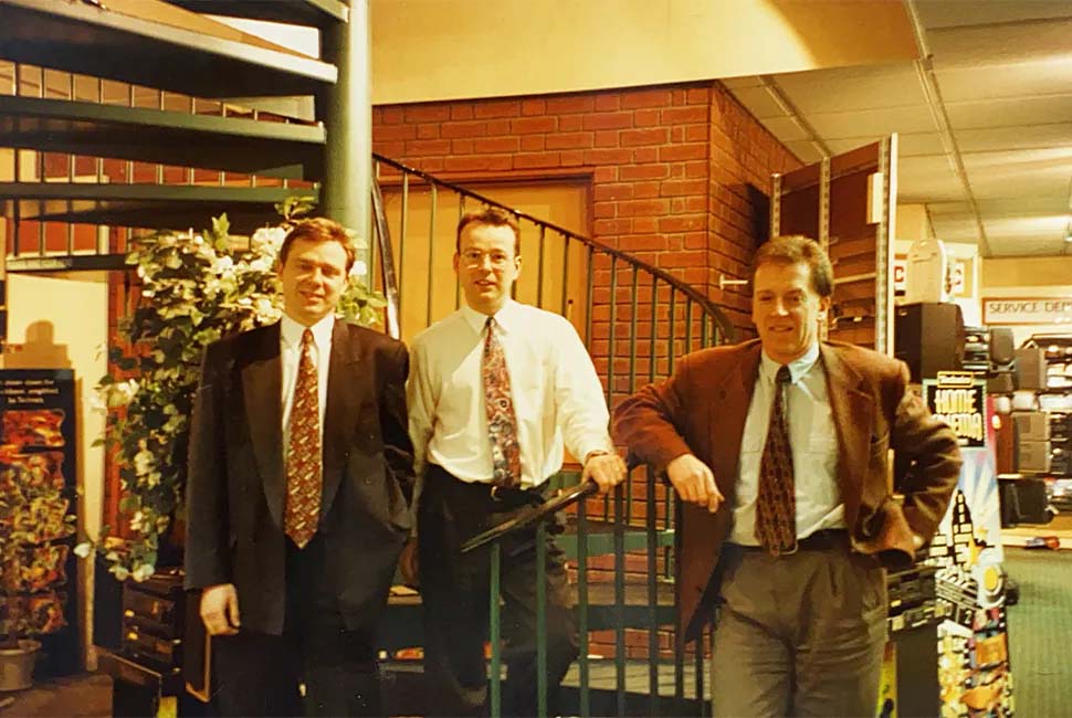 Three men in suits stand in front of a spiral staircase in an office setting, with green plants and furniture around, conveying a professional, relaxed atmosphere.
