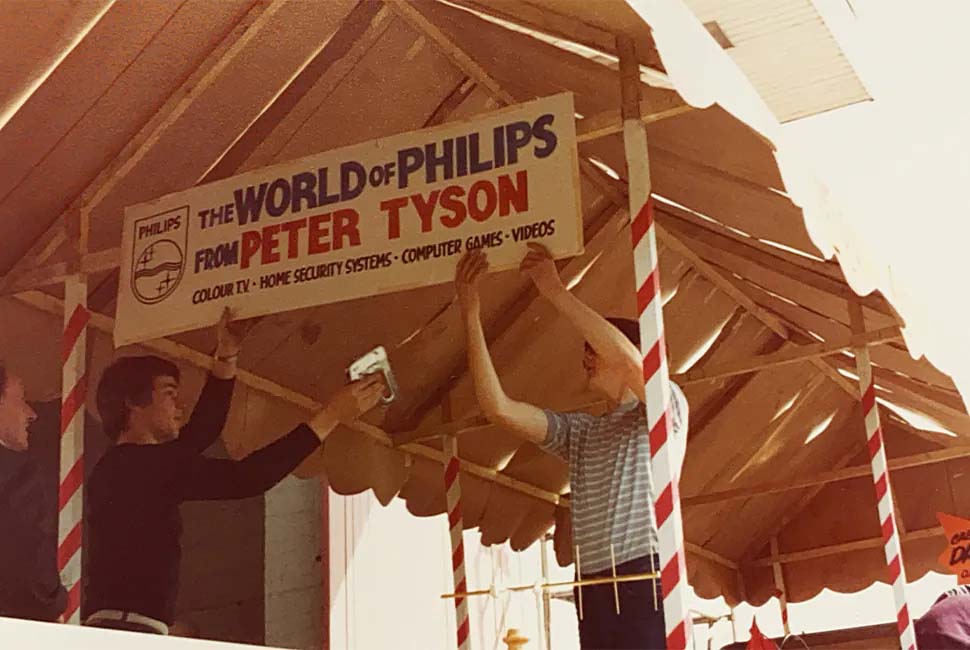 Three men work together to hang a sign under a striped awning. The sign reads "The World of Philips from Peter Tyson," promoting electronics and games.