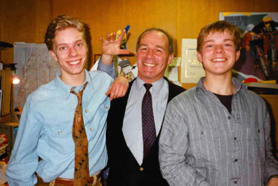 Three men smiling in an office setting. The man on the left gestures playfully, and the one in the center wears a suit. The mood is cheerful and relaxed.