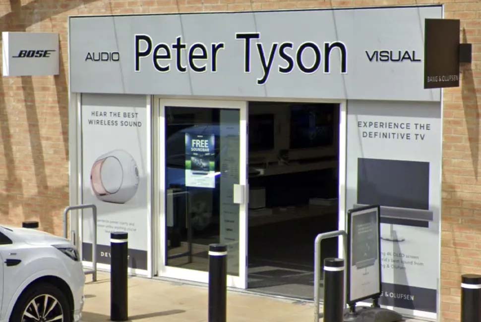 Storefront of "Peter Tyson Audio Visual," featuring large windows and open entrance. Prominent ads for wireless sound and TVs evoke modern tech vibes.