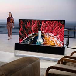 A modern living room with a large TV displaying a vivid image of red trees and a waterfall. A person stands by a railing in the background at sunset.