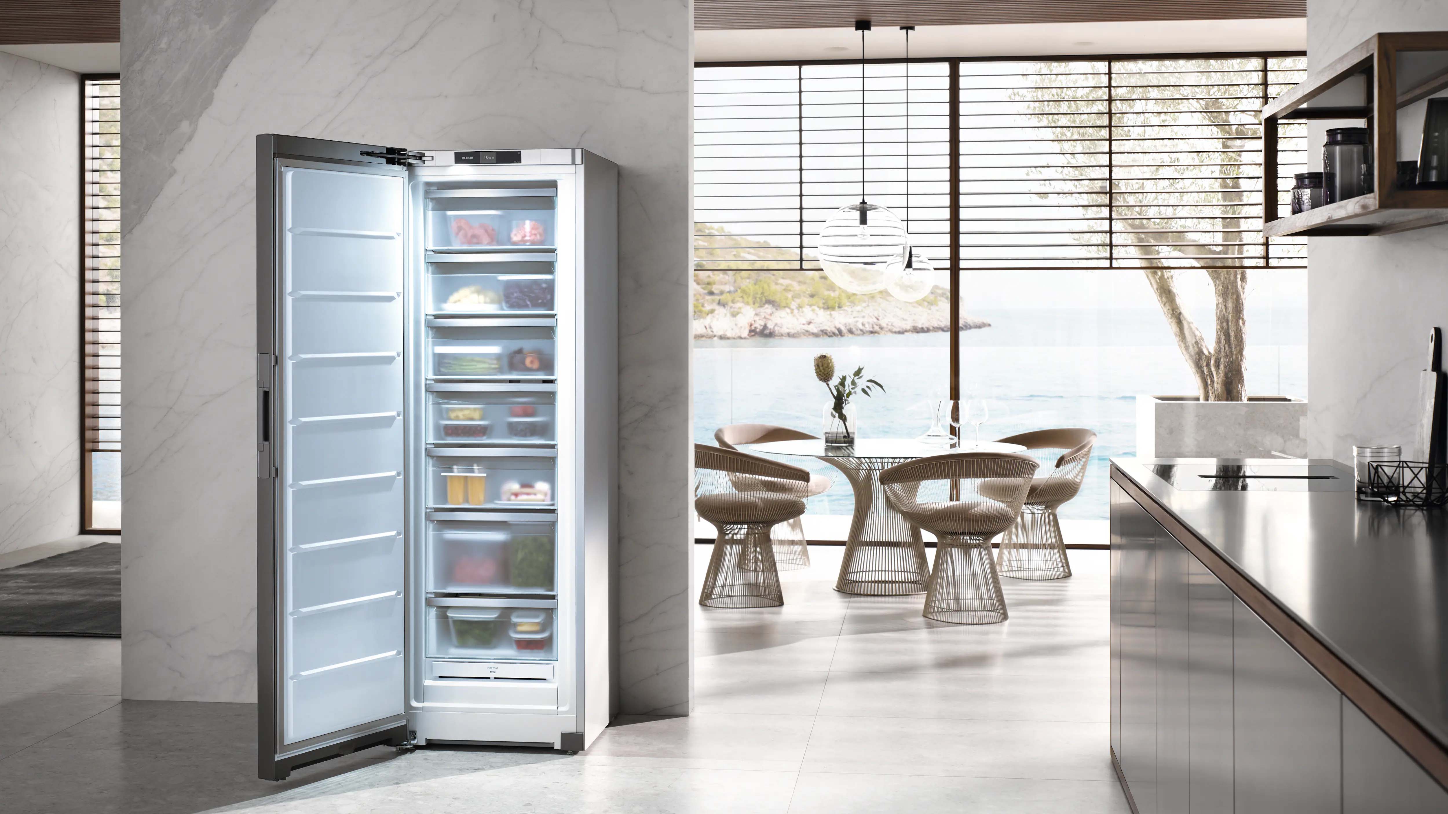 Modern kitchen with a stainless steel refrigerator open, showing organized shelves with food. Sunlit dining area with wicker chairs and sea view.