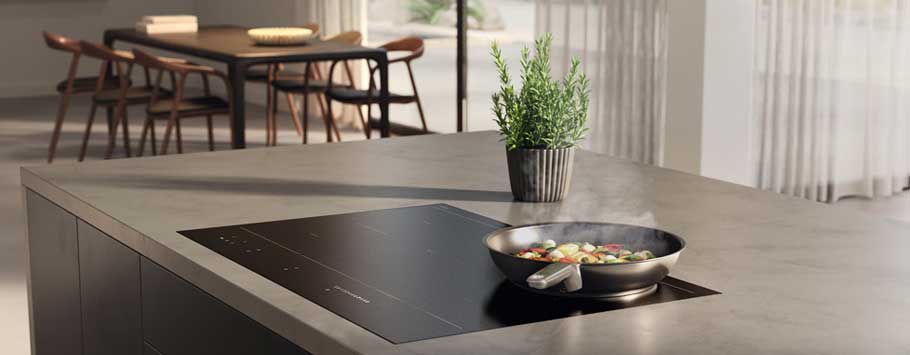 Modern kitchen with sleek gray countertop, black induction cooktop, and steaming pan of vegetables. Nearby, a small potted plant adds greenery.