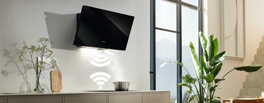 Modern kitchen with a sleek black wall-mounted range hood emitting light, casting shadows. A pot on the stove, plants, and minimal decor convey a calm vibe.