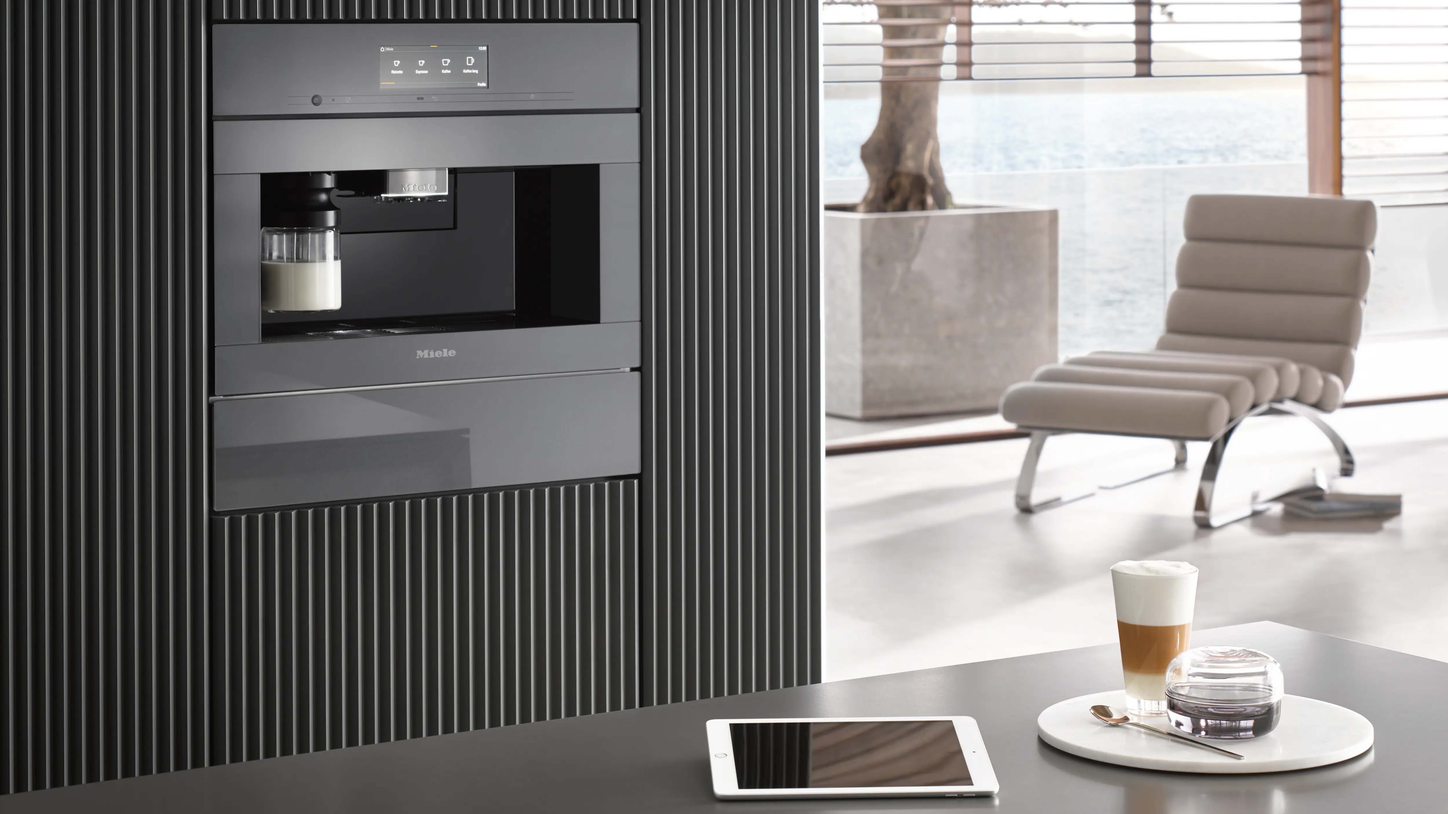 Modern kitchen with an integrated espresso machine in a sleek, black panel. A tablet, coffee, and dessert are on a counter. Soft light and a lounge chair create a relaxing atmosphere.