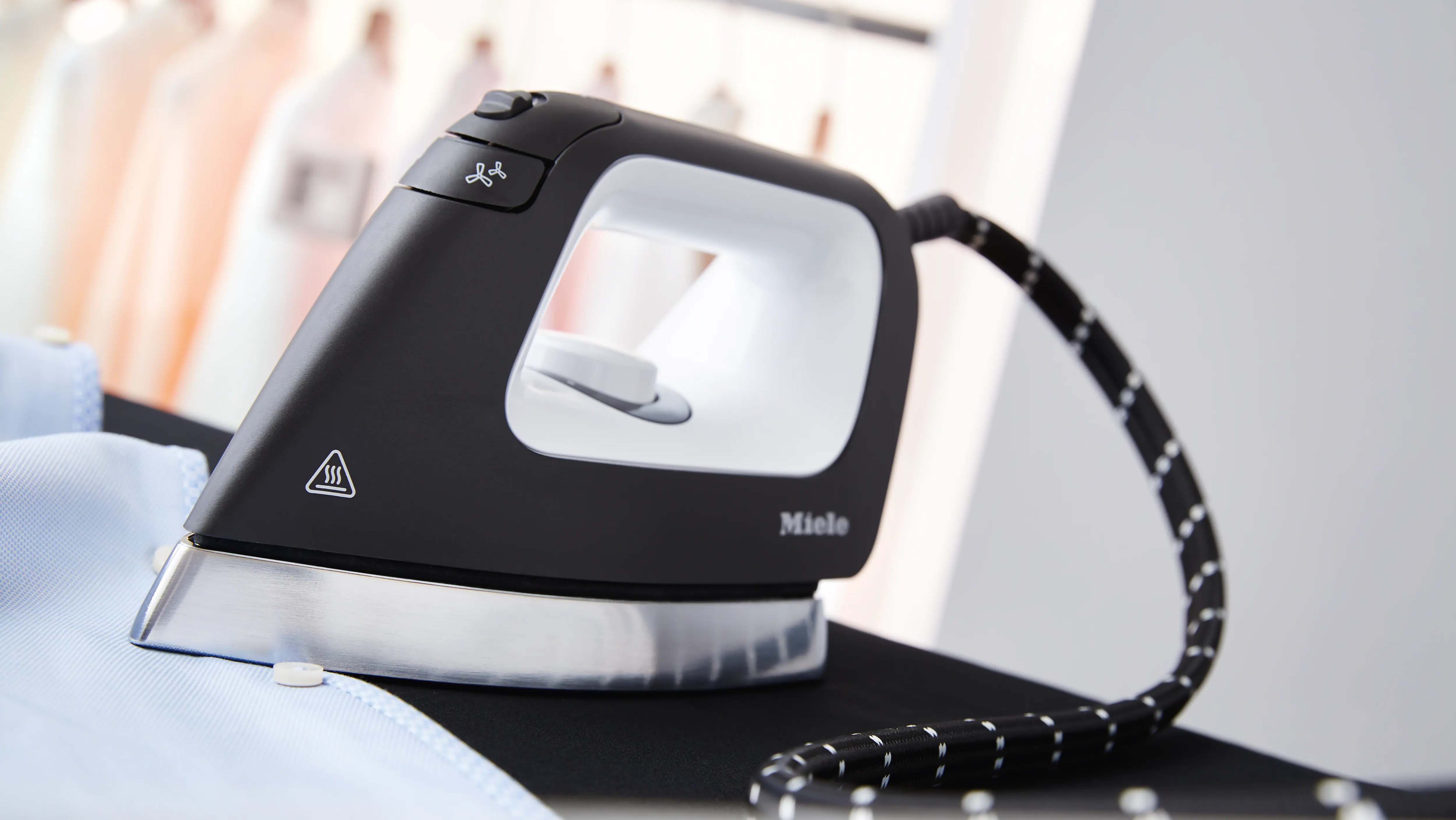 A black and white steam iron is positioned on a light blue garment. The iron's cord is visible, and blurred clothes hang in the background. The scene conveys a neat, organized atmosphere.