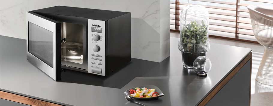 A sleek, modern microwave with its door open, revealing a clean interior. It sits on a kitchen counter beside a small plate of colorful fruit and a glass-domed plant, enhancing a minimalist and contemporary kitchen vibe.