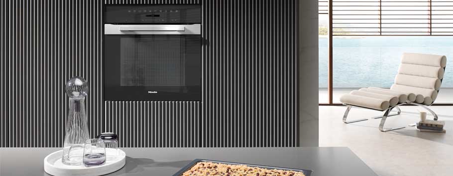 Modern kitchen interior with a black built-in oven set in a vertical striped wall. Foreground features a tray of cookies and glassware. A sleek lounge chair overlooks a serene waterfront view through large windows, infusing calm.