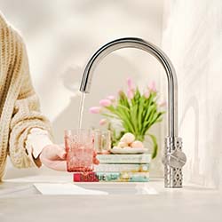 A person fills a pink glass with water from a sleek, silver kitchen faucet. In the background, blurred pink flowers and books create a cozy, fresh atmosphere.