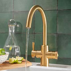Gold kitchen faucet running water into a sink, set against green tiled walls. A cutting board with limes and a glass carafe is nearby, creating a fresh, modern feel.