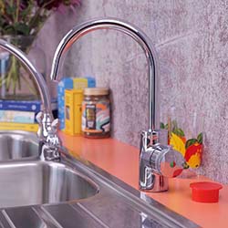 Chrome kitchen faucet on an orange countertop with a colorful glass, jars, and book in the background. The setting is clean and vibrant.